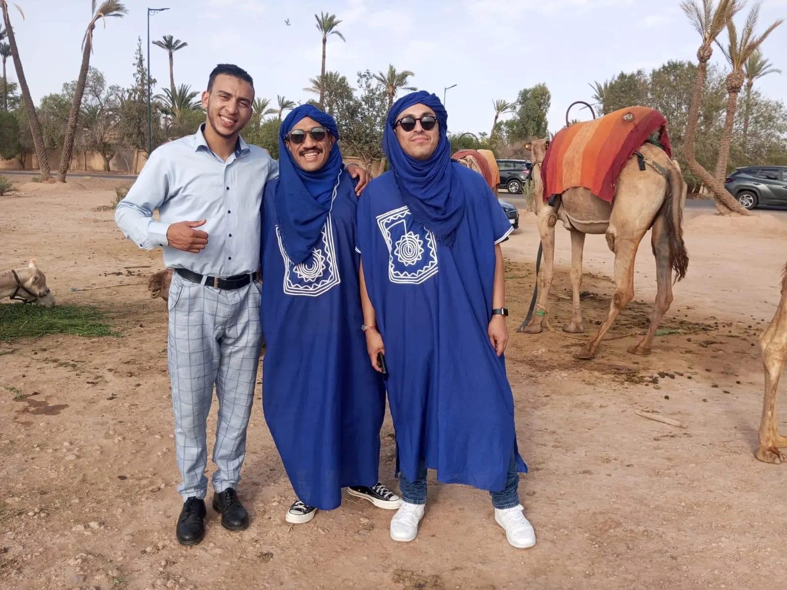 Deux clients en tenue touarègue bleue posant avec leur chauffeur dans la palmeraie de Marrakech, un dromadaire sellé derrière eux